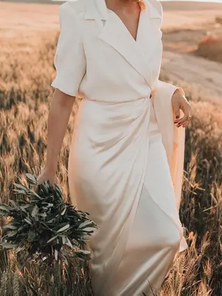 Novia con vestido cruzado de satén color marfil y ramo de olivo caminando por un campo de trigo al atardecer