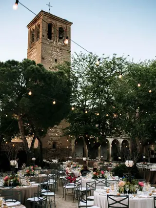 Banquete de boda al aire libre con mesas redondas, guirnaldas de luces y una torre de ladrillo histórica al fondo