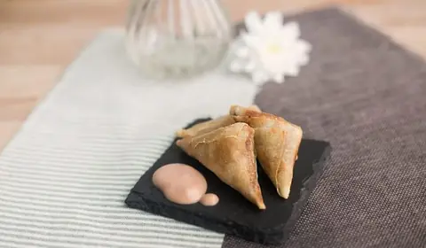 Tres triángulos de pasta crujiente dorada con una lágrima de salsa rosada sobre pizarra, junto a un jarrón y una margarita