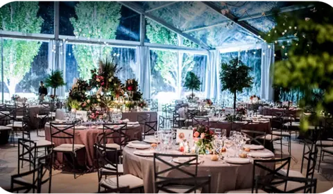 Carpa transparente iluminada por la noche con mesas redondas de mantel rosado, sillas negras y centros de flores altos