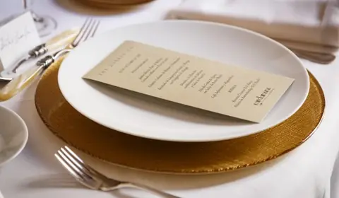 Minuta impresa sobre un plato blanco y bajoplato de cristal dorado, con cubiertos de plata y tarjeta de sitio en la mesa