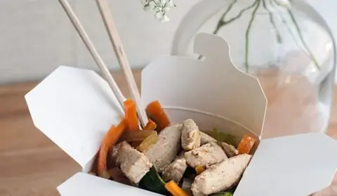 Caja de cartón blanca abierta con dados de pollo salteados con zanahoria y calabacín, y unos palillos de madera