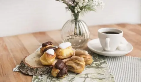 Petisús rellenos de crema con cobertura de chocolate y glaseado blanco sobre una rodaja de tronco junto a una taza de café