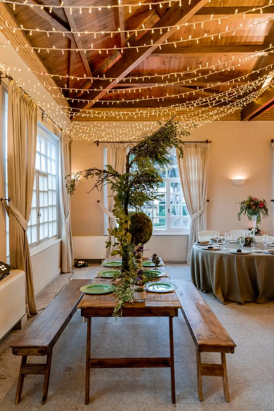 Mesa de madera con bancos, vajilla verde y centro de ramas bajo guirnaldas de luces en un salón de eventos