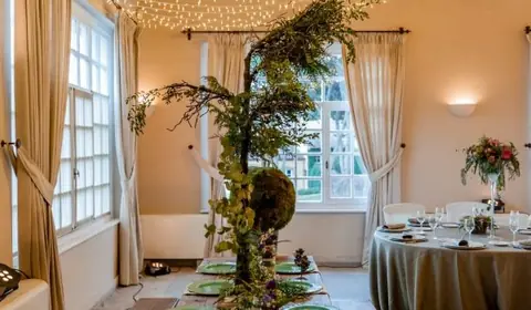Mesa de madera con bancos, vajilla verde y centro de ramas bajo un techo de guirnaldas de luces en un salón