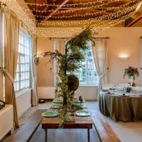 Mesa de madera con bancos, vajilla verde y centro de ramas bajo un techo de guirnaldas de luces en un salón