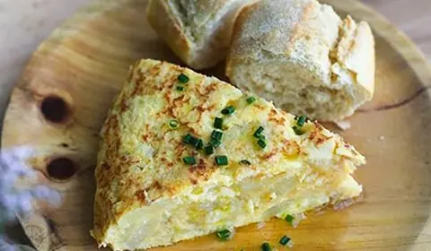 Porción de tortilla de patata jugosa con cebollino picado y dos trozos de pan sobre un plato de madera