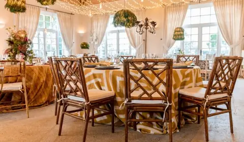 Salón con mesas redondas de mantel estampado en dorado y blanco, sillas de bambú, guirnaldas de luces y lámparas de plantas