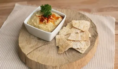 Cuenco cuadrado de hummus con pimentón, aceite y perejil junto a crujientes de pan sobre una rodaja de tronco
