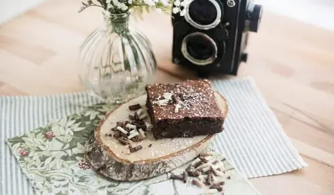 Porción de brownie de chocolate con virutas de chocolate y coco rallado sobre una rodaja de tronco, con una cámara antigua