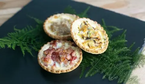 Tres mini quiches de masa quebrada, una con beicon, otra con cebolla y otra con verduras, sobre pizarra con hojas de helecho