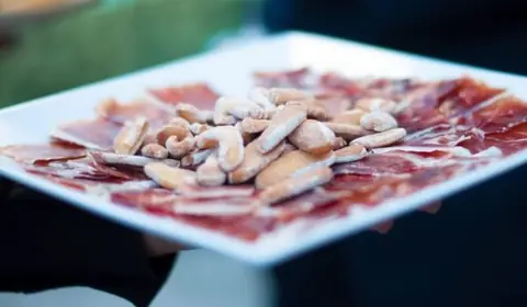 Bandeja blanca con lonchas finas de jamón ibérico y picos de pan en el centro, sostenida por un camarero en un evento
