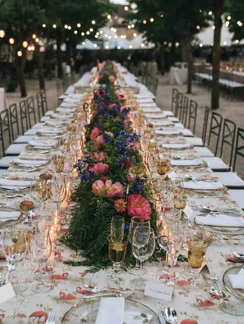 Larga mesa imperial de boda con camino de peonías, delphinium y verde, velas y mantel estampado de flamencos