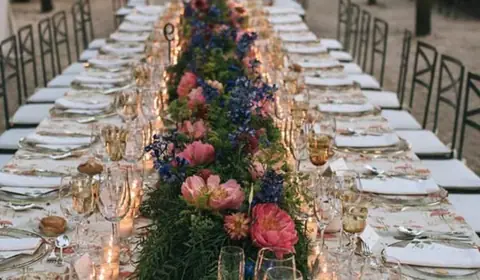 Larga mesa imperial de boda con camino de peonías, delphinium y verde, velas y mantel estampado de flamencos