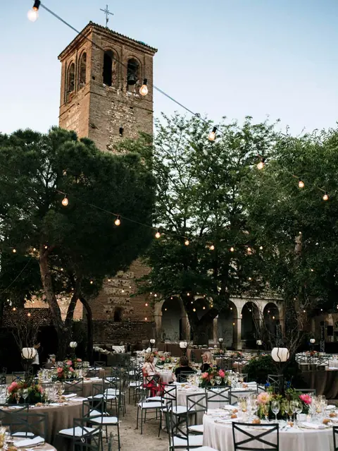 Banquete de boda al aire libre con mesas redondas, guirnaldas de luces y una torre de ladrillo histórica al fondo