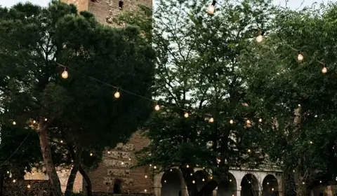 Banquete de boda al aire libre con mesas redondas, guirnaldas de luces y una torre de ladrillo histórica al fondo