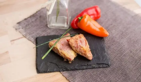 Dos bocados empanados de interior rojizo sobre pizarra con cebollino, junto a dos pimientos dulces rojo y naranja