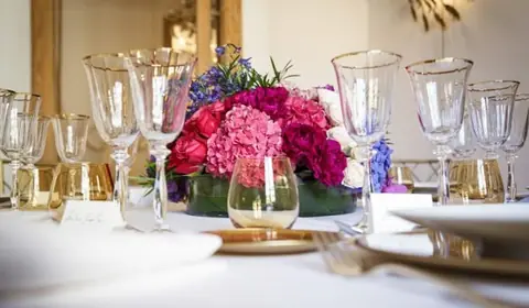 Mesa vestida de blanco con centro bajo de hortensias, rosas y peonías fucsias, copas con filo dorado y tarjetas de sitio