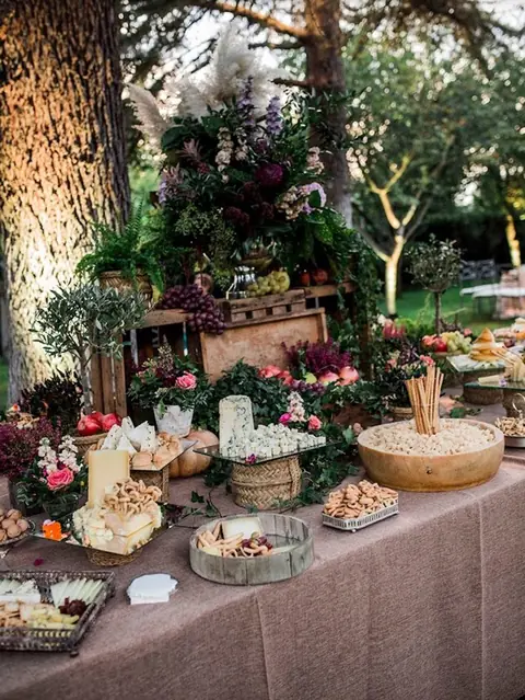 Bufé de quesos con tablas, panes, colines y cajas de madera decoradas con flores bajo los árboles en una boda