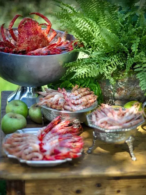 Mesa de marisco con centollo, carabineros y gambas en fuentes de plata junto a helechos y manzanas en un evento