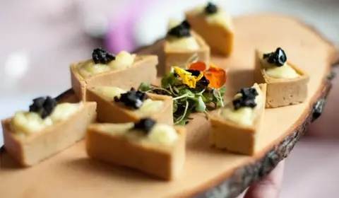 Tartaletas cuadradas rellenas de crema y coronadas con un toque oscuro sobre una tabla de tronco con flores comestibles