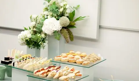 Expositor de cristal con mini sándwiches, montaditos y sushi junto a un gran arreglo de hortensias blancas en una oficina