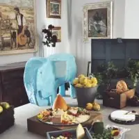 Mesa de catering con quesos, pan, fruta, girasoles y una escultura de elefante en un salón con cuadros