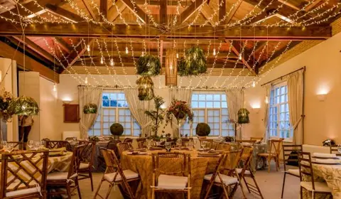 Salón con vigas de madera, guirnaldas de luces, lámparas de plantas y mesas redondas montadas con sillas de bambú