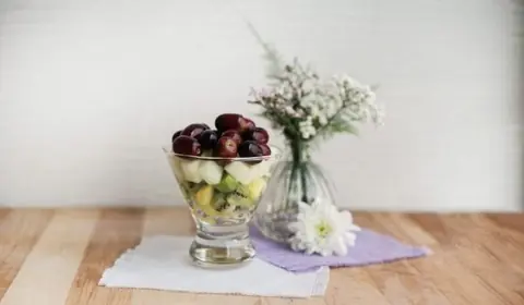 Copa de cristal con macedonia de kiwi, pera y piña coronada con uvas negras, junto a un jarrón de flores