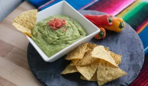Cuenco de guacamole con tomate picado y nachos de maíz sobre pizarra redonda, con pimientos dulces y una manta de colores