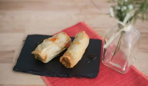 Dos rollitos de primavera dorados con salsa agridulce sobre una pizarra, con mantel rojo y un pequeño jarrón de cristal