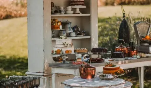 Aparador blanco antiguo con tartas, teteras y tazas en un jardín, junto a una mesa de cobre y un carrito de copas