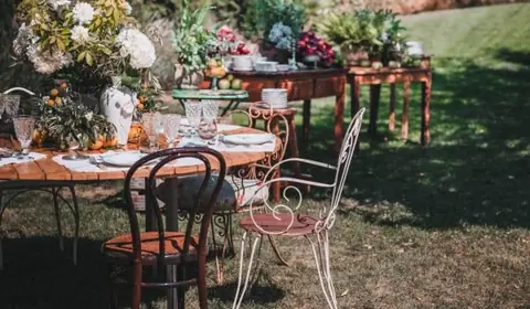 Mesa de boda en un jardín con centro de hortensias y flores blancas, naranjas, copas y sillas de madera y forja
