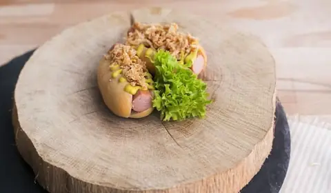 Mini hot dogs con salchicha, mostaza y cebolla crujiente junto a una hoja de lechuga rizada sobre una rodaja de tronco