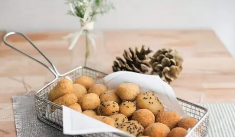 Cesta metálica de freír con croquetitas variadas, algunas con sésamo negro, sobre papel, junto a dos piñas secas