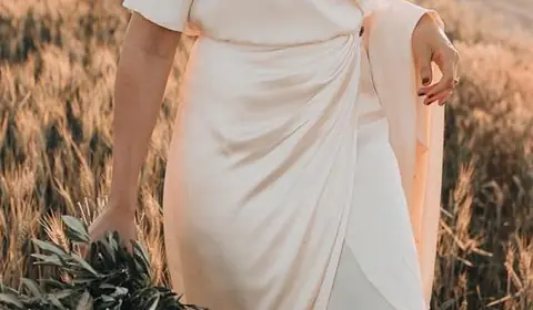 Novia con vestido cruzado de satén color marfil y ramo de olivo caminando por un campo de trigo al atardecer