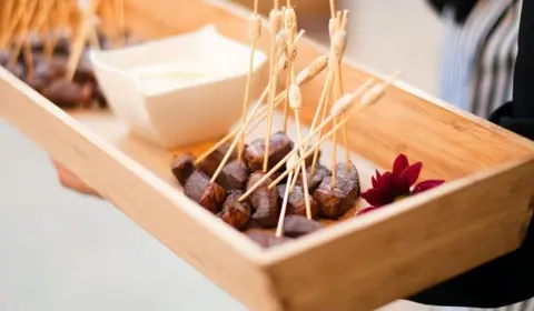 Dados de carne glaseada en palillos de bambú dentro de una bandeja de madera con salsa y una dalia roja