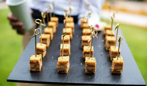 Pizarra alargada con bocados de pan de especias y foie pinchados en palillos, llevada por un camarero en un jardín