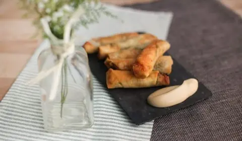 Rollitos alargados de pasta crujiente y dorada sobre una pizarra con una lágrima de salsa, junto a un jarrón con lazo