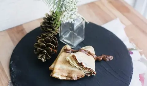 Dos crepes doblados en triángulo con crema de chocolate sobre una pizarra redonda, junto a piñas y un jarrón de flores