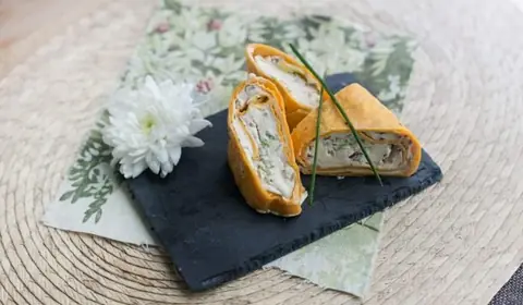 Wraps de tortilla anaranjada cortados por la mitad con queso crema, pollo y verduras sobre pizarra y servilleta floral