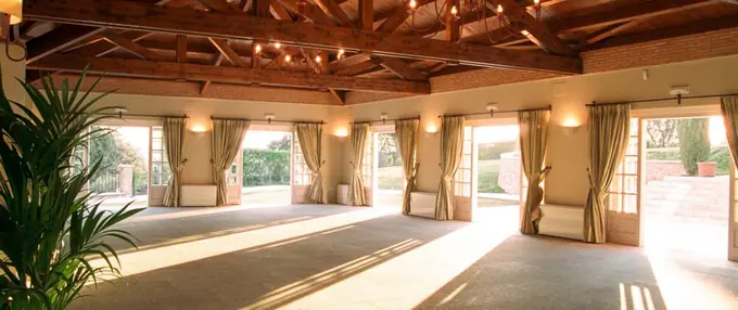 Un amplio salón con techo de vigas de madera a la vista, iluminado por grandes ventanales y lámparas de araña, ideal para eventos. Las cortinas pesadas adornan las puertas de vidrio que se abren hacia el exterior, complementando el ambiente rústico y elegante del lugar. Plantas en macetas añaden un toque natural al espacio.