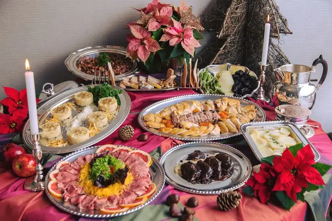 una mesa festiva repleta de diferentes platillos dispuestos elegantemente en bandejas de plata. En el centro, hay un arreglo floral de nochebuenas, que junto a las piñas y las velas encendidas, sugieren que se trata de una cena navideña o de fin de año. Los platillos incluyen lo que parece ser un asado de carne frío adornado con hierbas y rodajas de naranja, canapés, verduras al vapor, y posiblemente algunos platillos de inspiración escandinava como arenques o salmón. El fondo de la imagen es neutro, lo que permite que el colorido de la comida y la decoración resalten, creando una atmósfera cálida y acogedora.