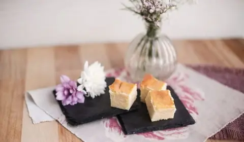 Dados de tarta de queso dorada por encima sobre dos pizarras, con un crisantemo blanco, otro malva y un jarrón de flores