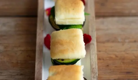 Mini focaccias rellenas de calabacín a la plancha alineadas en una bandeja estrecha de madera con pétalos de rosa
