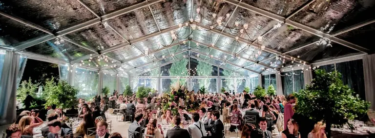 Banquete de boda bajo una carpa transparente con lámparas de araña, árboles y cientos de invitados cenando
