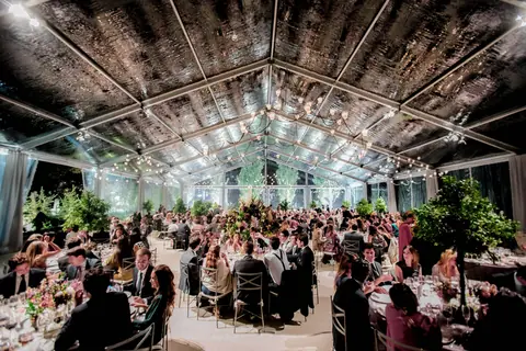 Banquete de boda bajo una carpa transparente con lámparas de araña, árboles y cientos de invitados cenando