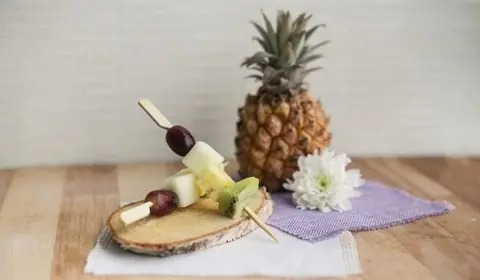 Brocheta de uvas, melón, piña y kiwi sobre una rodaja de tronco, con una piña entera y un crisantemo blanco detrás