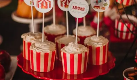 Cupcakes en cápsulas de rayas rojas y blancas con toppers de circo sobre un soporte rojo en una mesa dulce