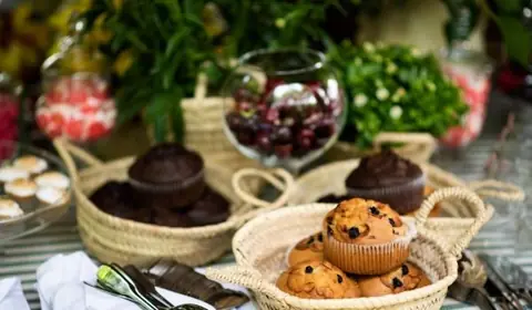 Capazos de palma con magdalenas de pepitas de chocolate y de chocolate, una copa de uvas y plantas sobre mantel de rayas